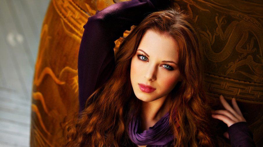 A woman with long red hair and blue eyes is wearing a purple scarf.