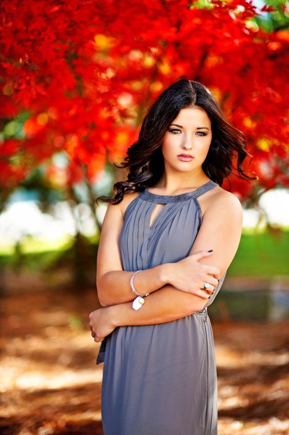 A woman in a grey dress is standing in front of a tree with red leaves.