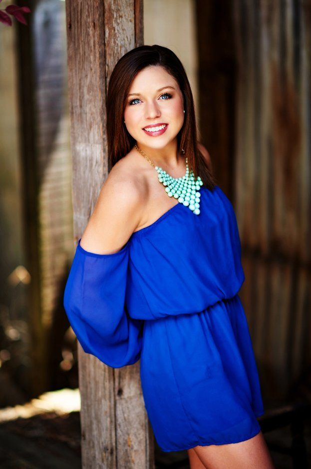 A woman in a blue dress is leaning against a wooden post.