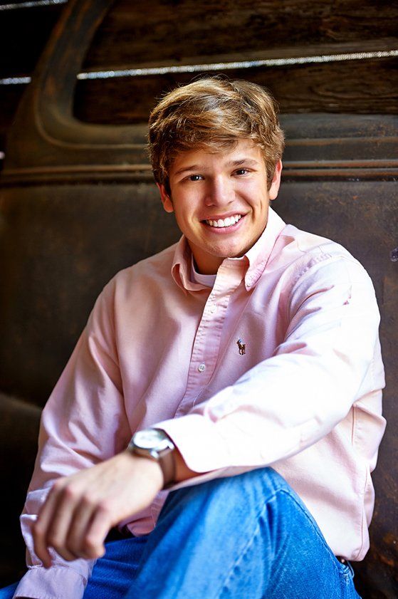 A young man in a pink shirt and blue jeans is sitting on a bench.
