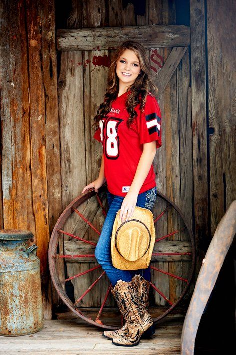 A woman in a red jersey and cowboy boots is standing next to a wagon wheel.