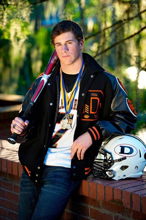 A young man is leaning against a brick wall holding a baseball bat and a football helmet.