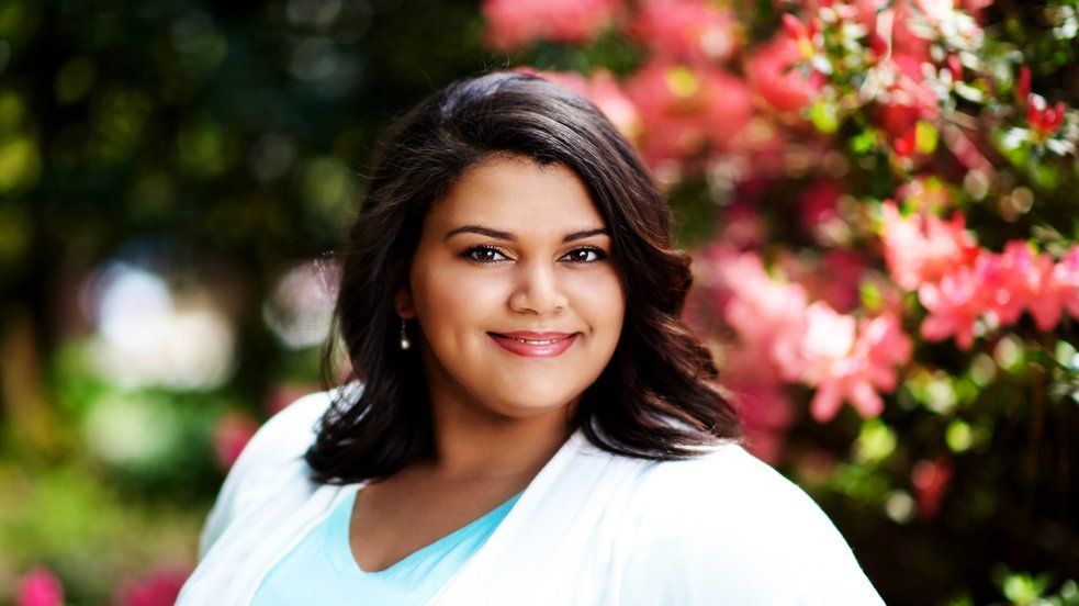 A woman is smiling in front of a bush of pink flowers.