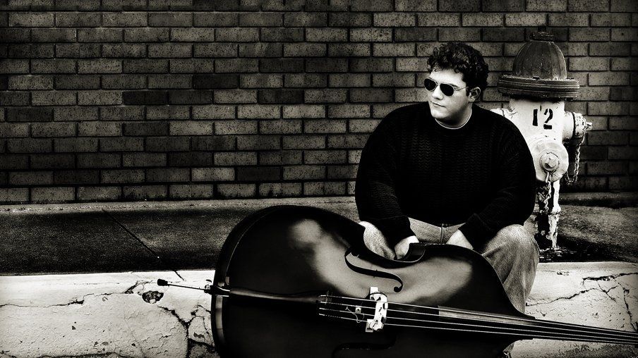A man is sitting on the sidewalk with a double bass in front of a fire hydrant with the number 12 on it