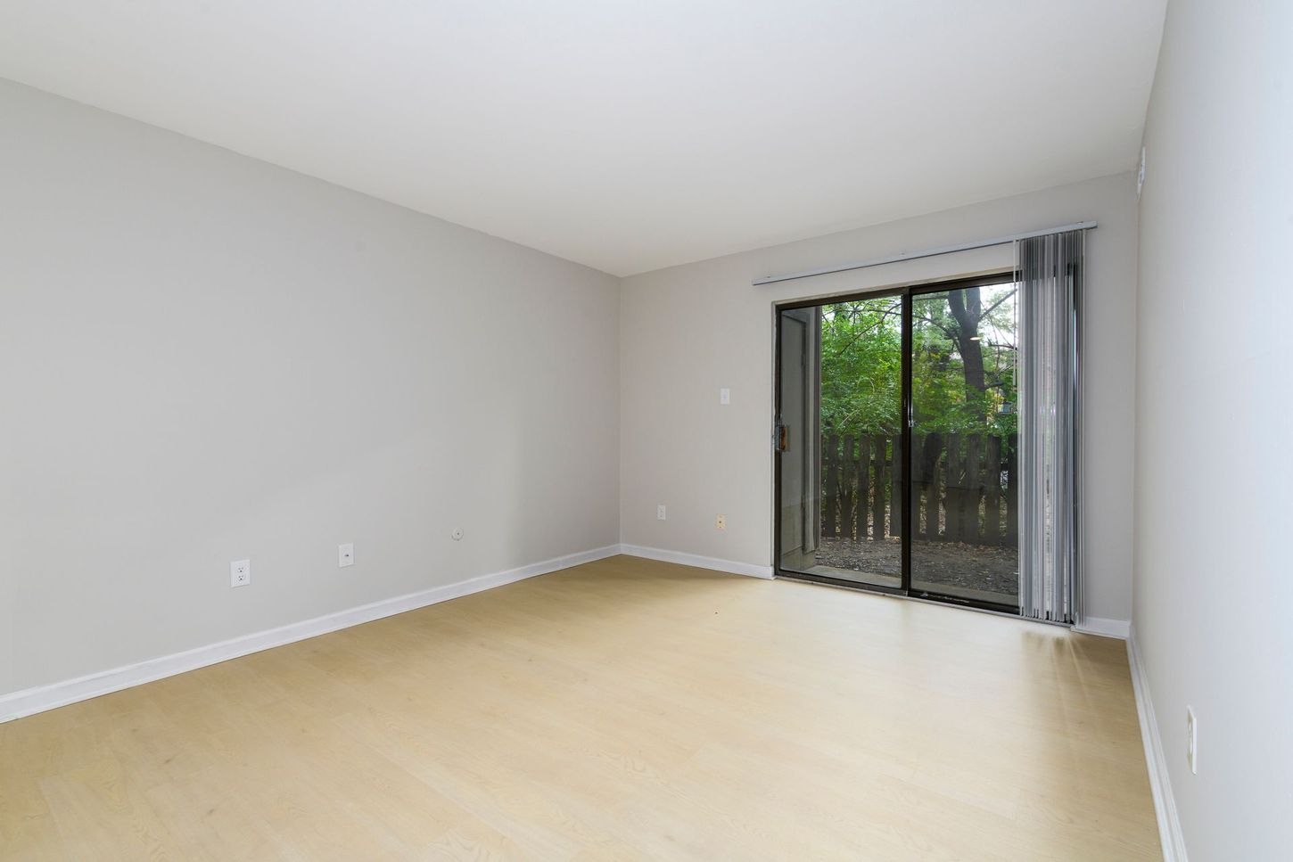 Empty room with light wood floors, gray walls, and a sliding glass door to a wooded area.