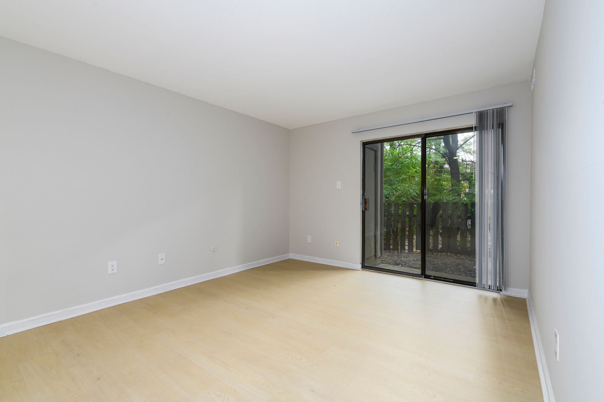 Empty room with light wood floors, gray walls, and a sliding glass door to a wooded area.