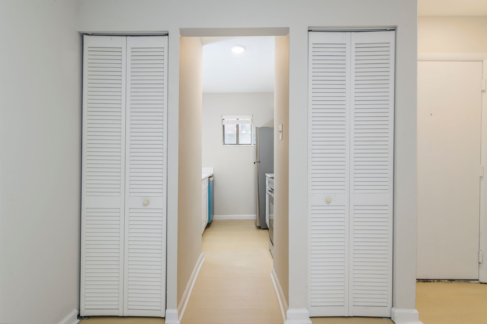 Hallway with white louvered closet doors on each side, leading to a kitchen with a window and appliances.