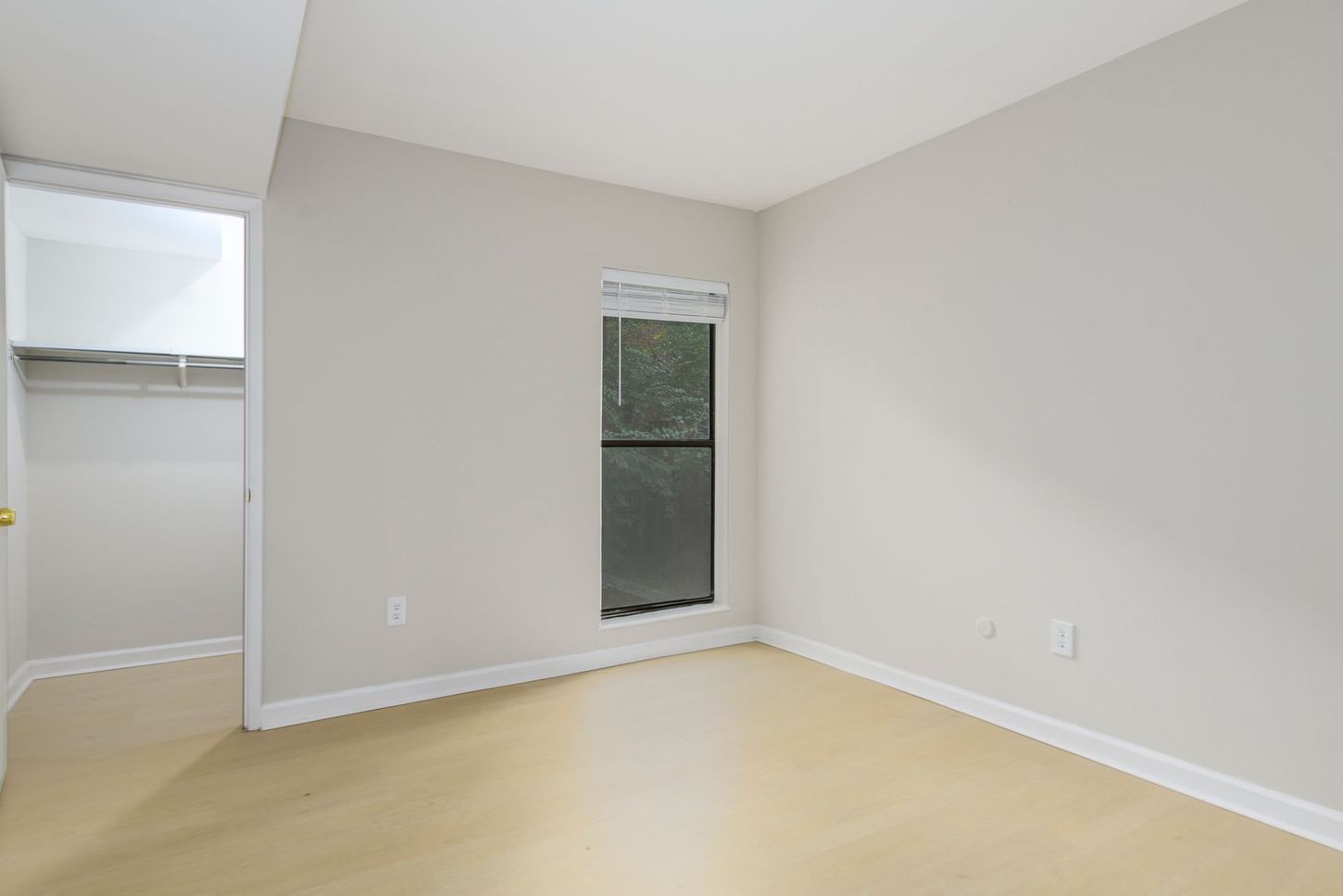 Empty bedroom with closet and window; beige walls, light wood floor.