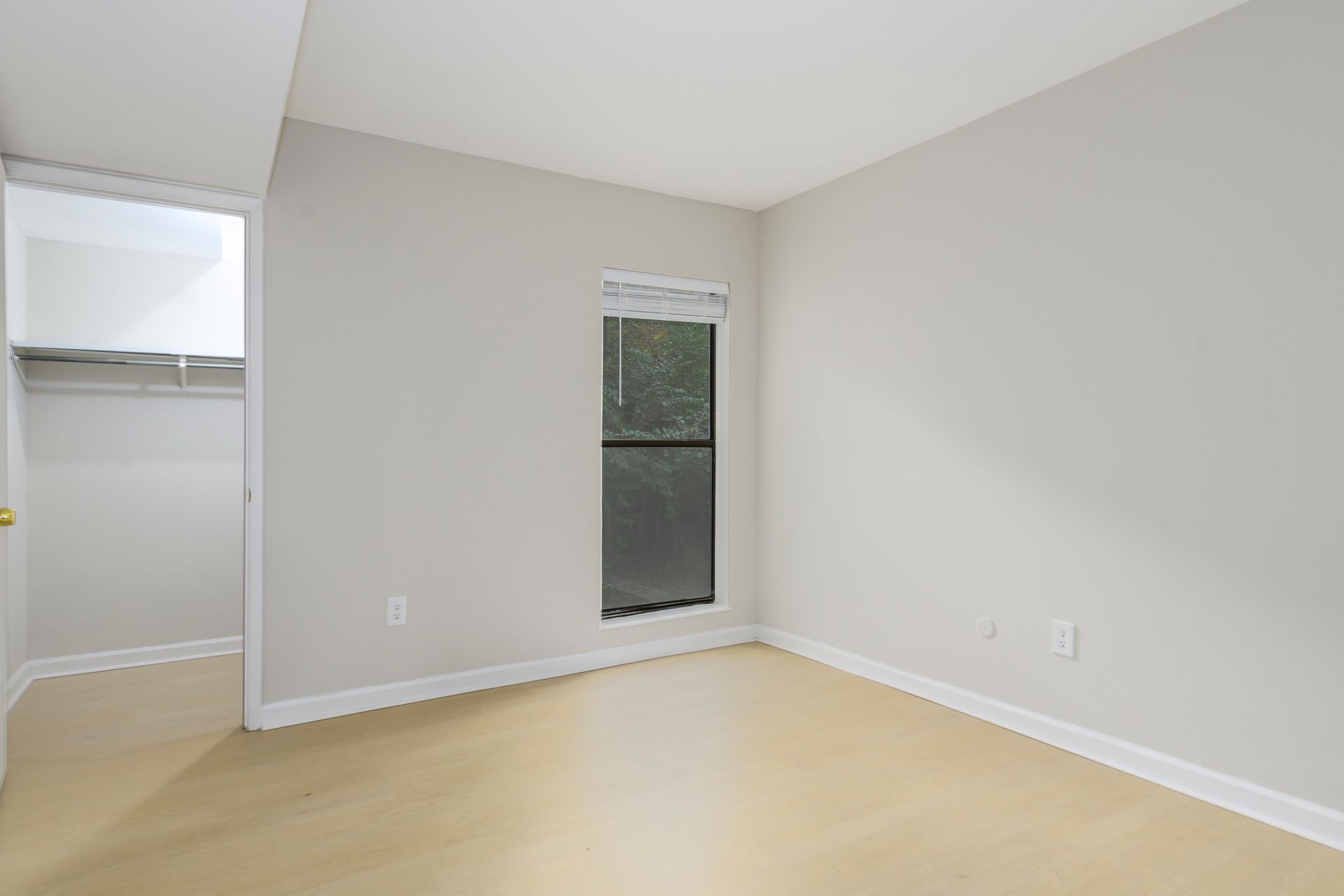 Empty bedroom with closet and window; beige walls, light wood floor.