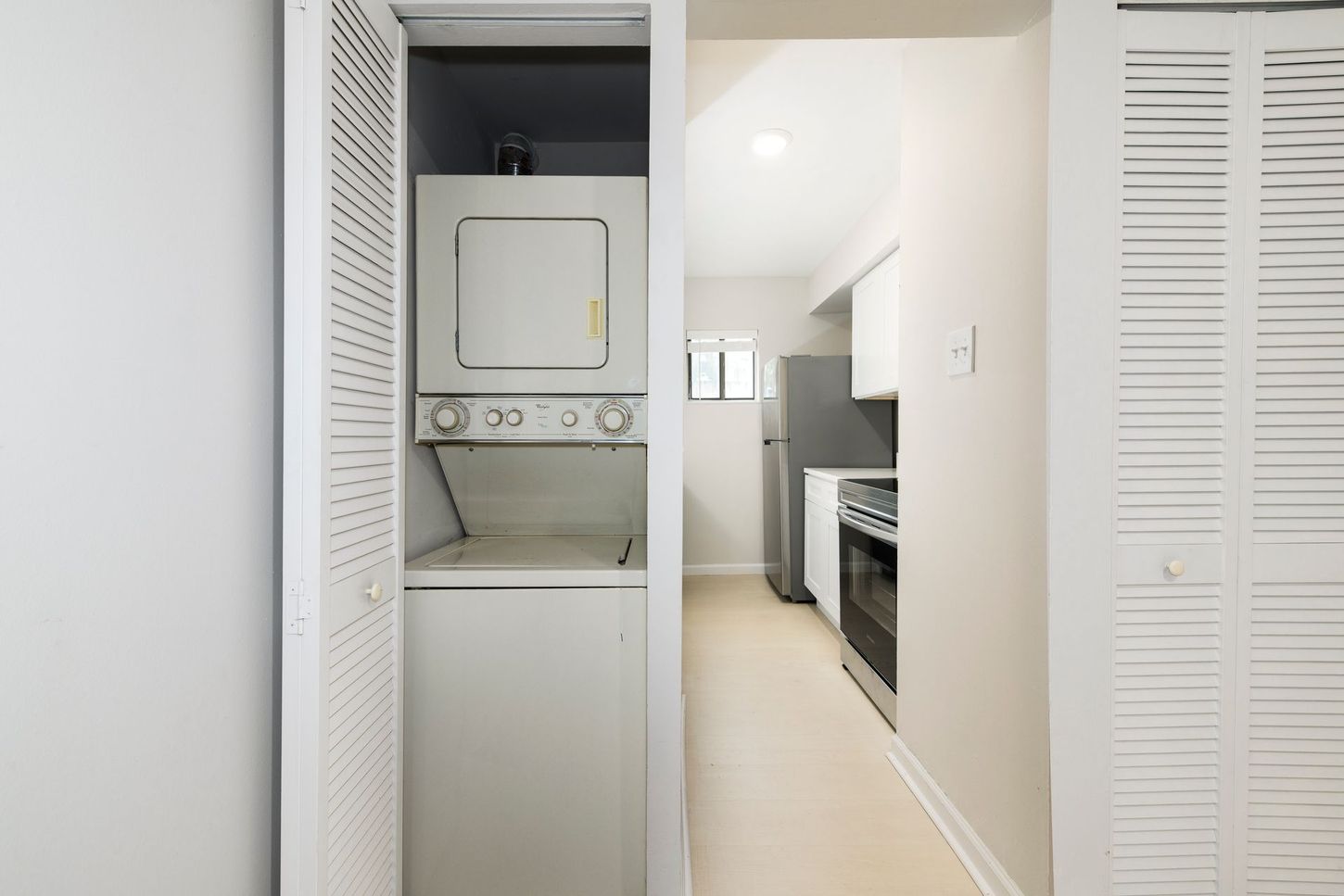 Stacked white washer and dryer in a narrow hallway with a kitchen visible in the background.