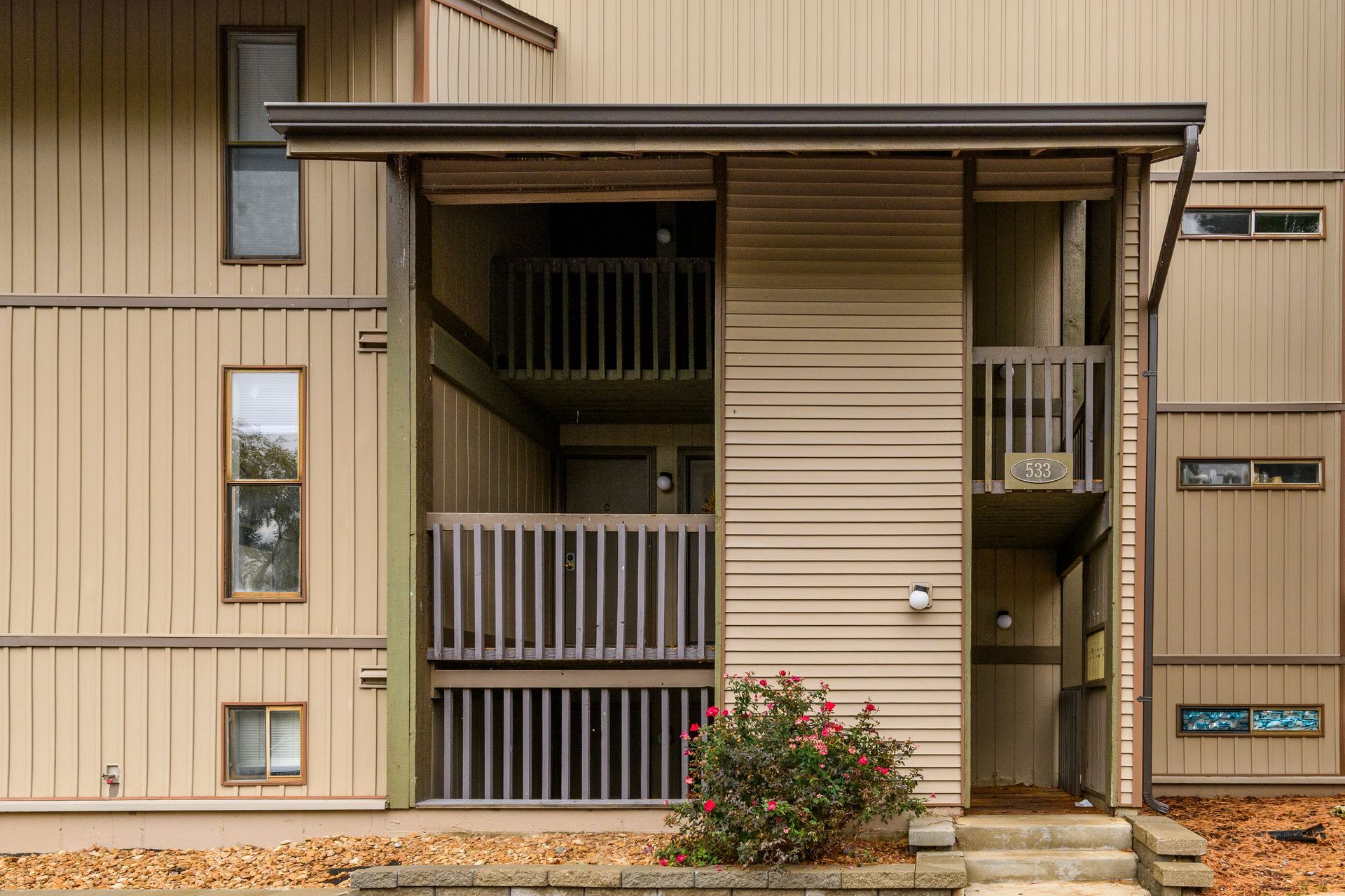 Tan and brown apartment building with balconies and a small rose bush.