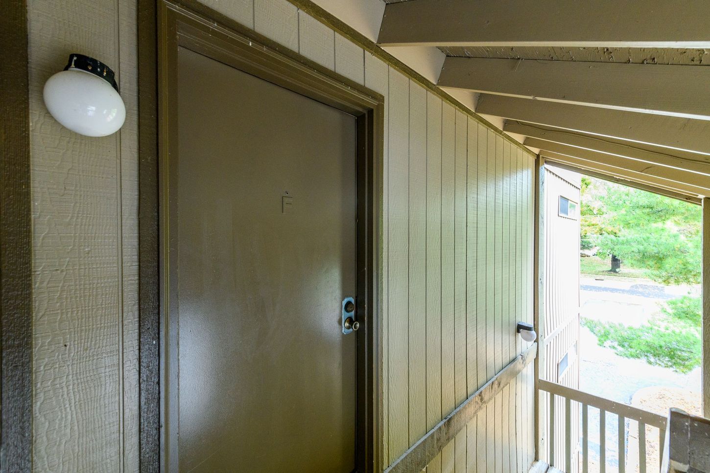 Brown door on a covered porch with a light fixture and railing, looking towards greenery.