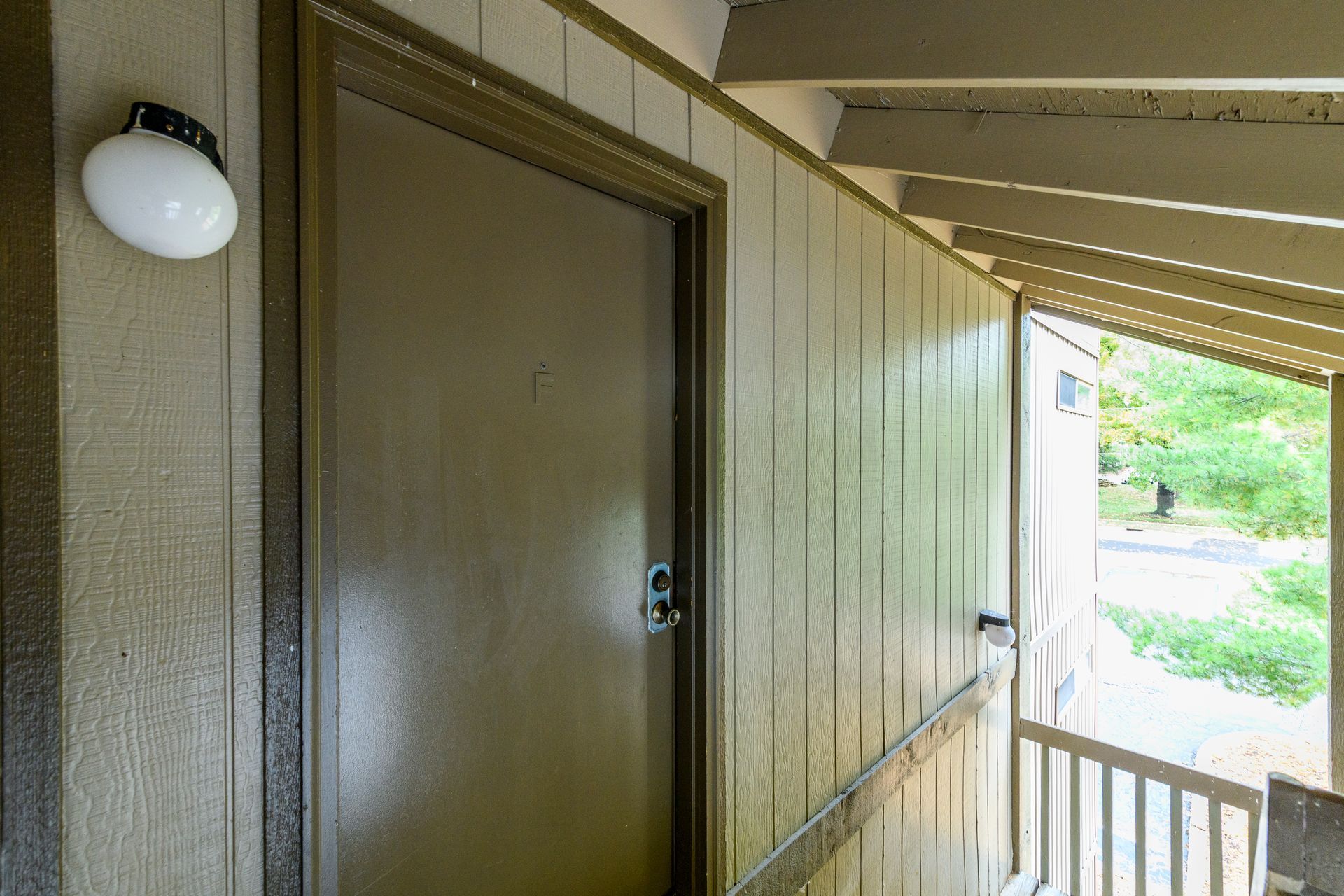 Brown door on a covered porch with a light fixture and railing, looking towards greenery.