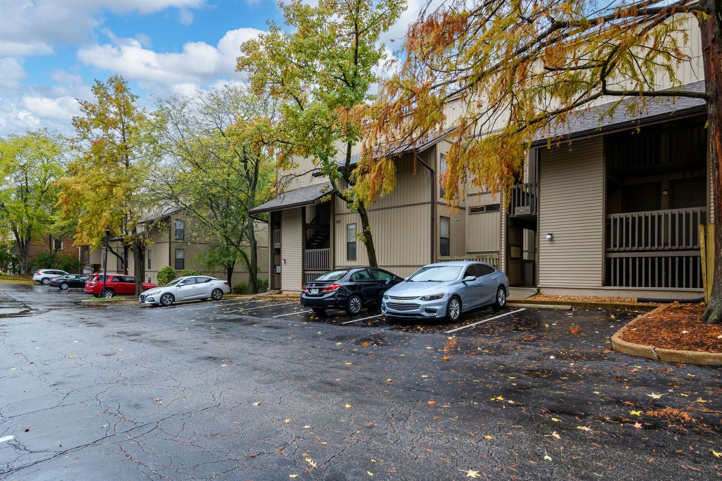 Apartment building exterior with parked cars on a wet parking lot, trees with fall foliage.