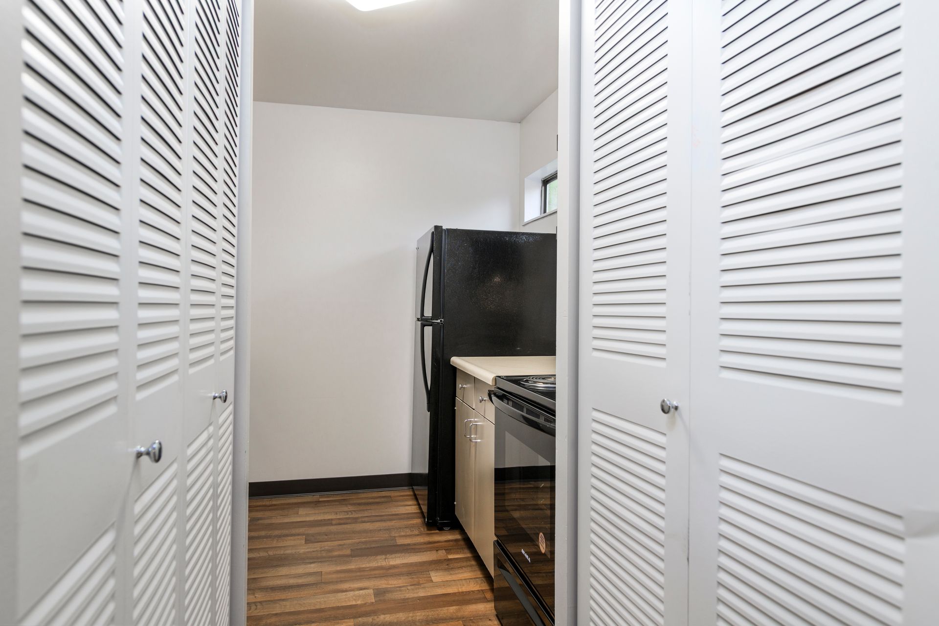 Narrow kitchen with white louvered closet doors, black refrigerator, and wood-look flooring.