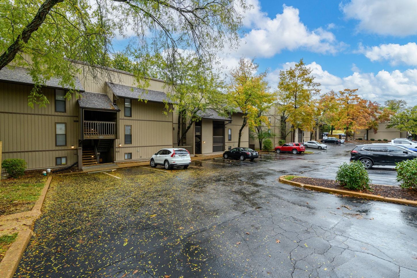 Apartment building with cars parked out front, trees, and cloudy sky.