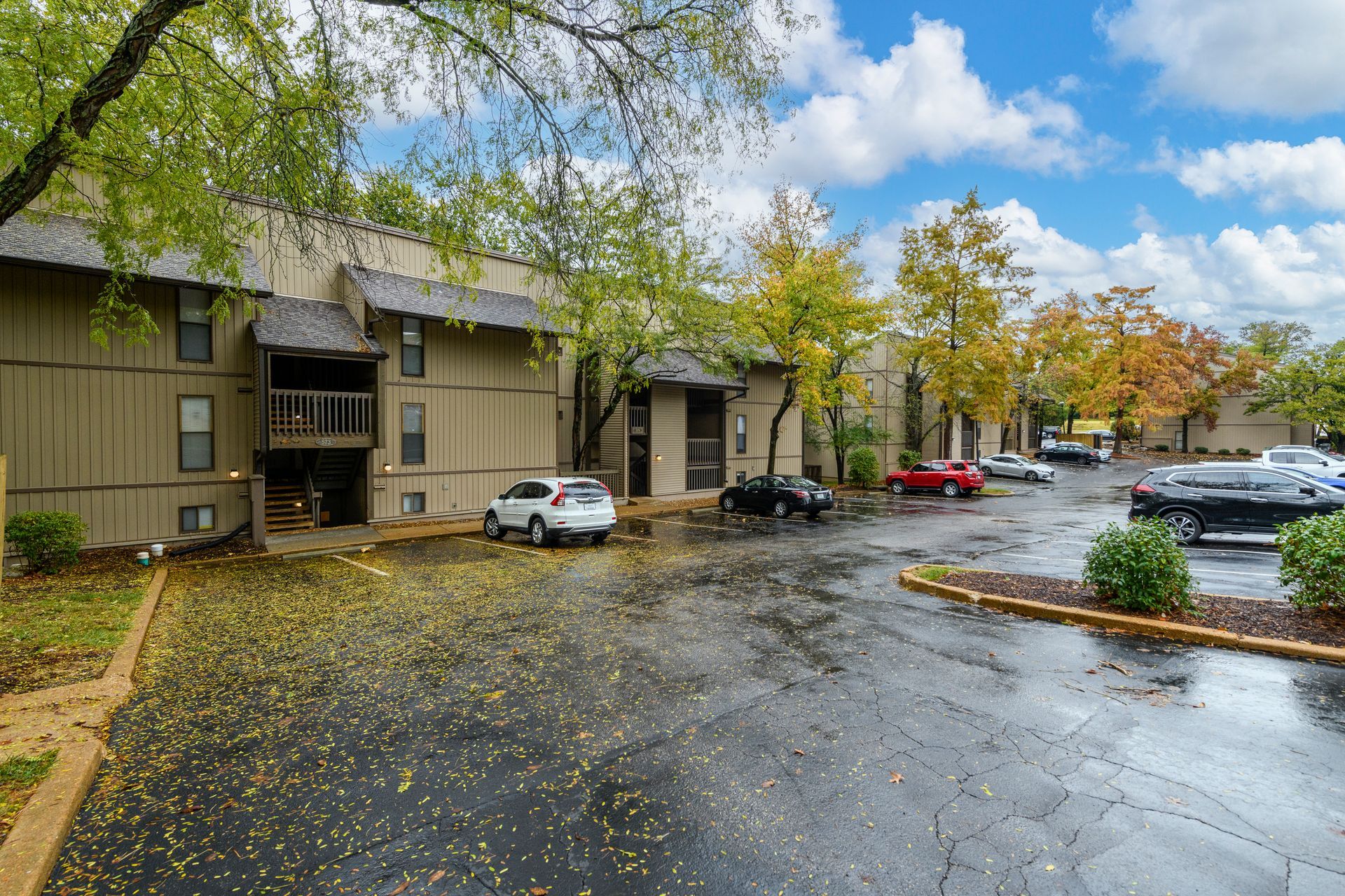Apartment building with cars parked out front, trees, and cloudy sky.