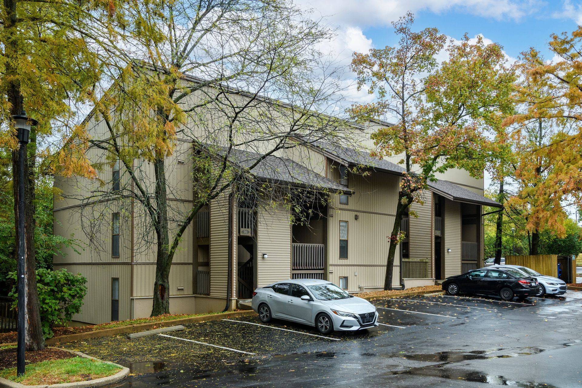 Apartment building exterior, beige siding, parked cars. Trees with fall foliage. Cloudy sky.