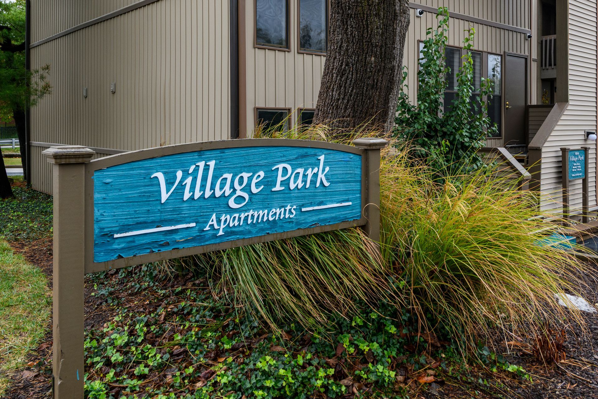 Sign for Village Park Apartments, teal background, brown frame, with a building in background.