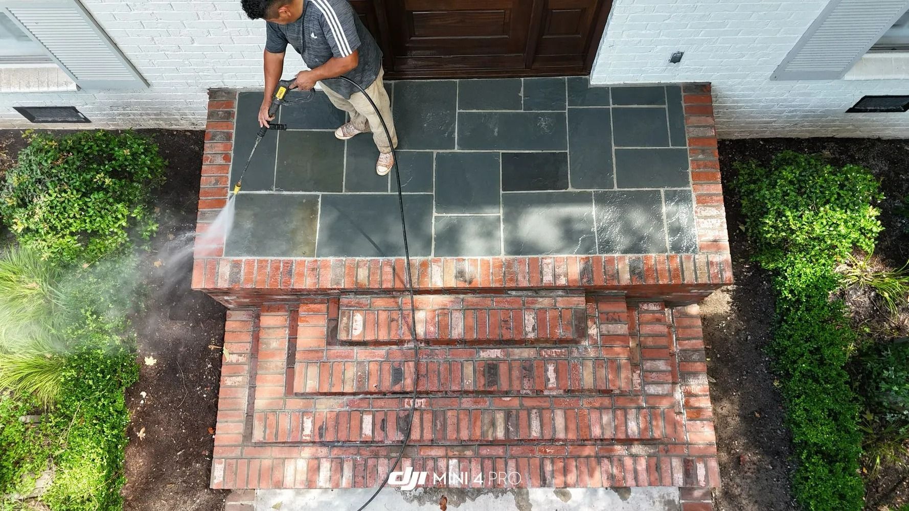 Worker on a rooftop repairing black shingles beside a brick chimney, viewed from above.