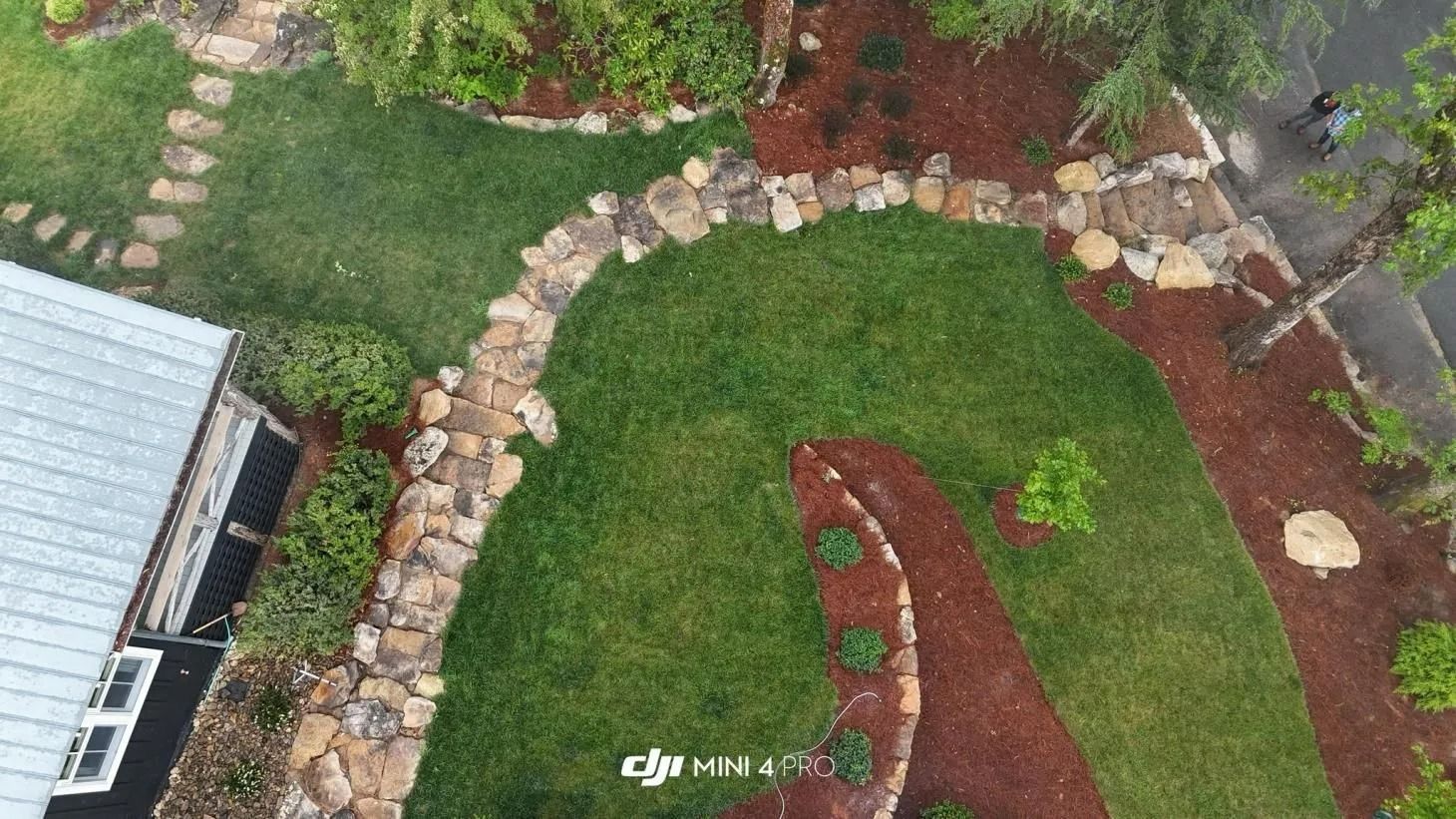 Aerial view of a landscaped backyard with green lawn, stone border, and red mulch beds beside a house.