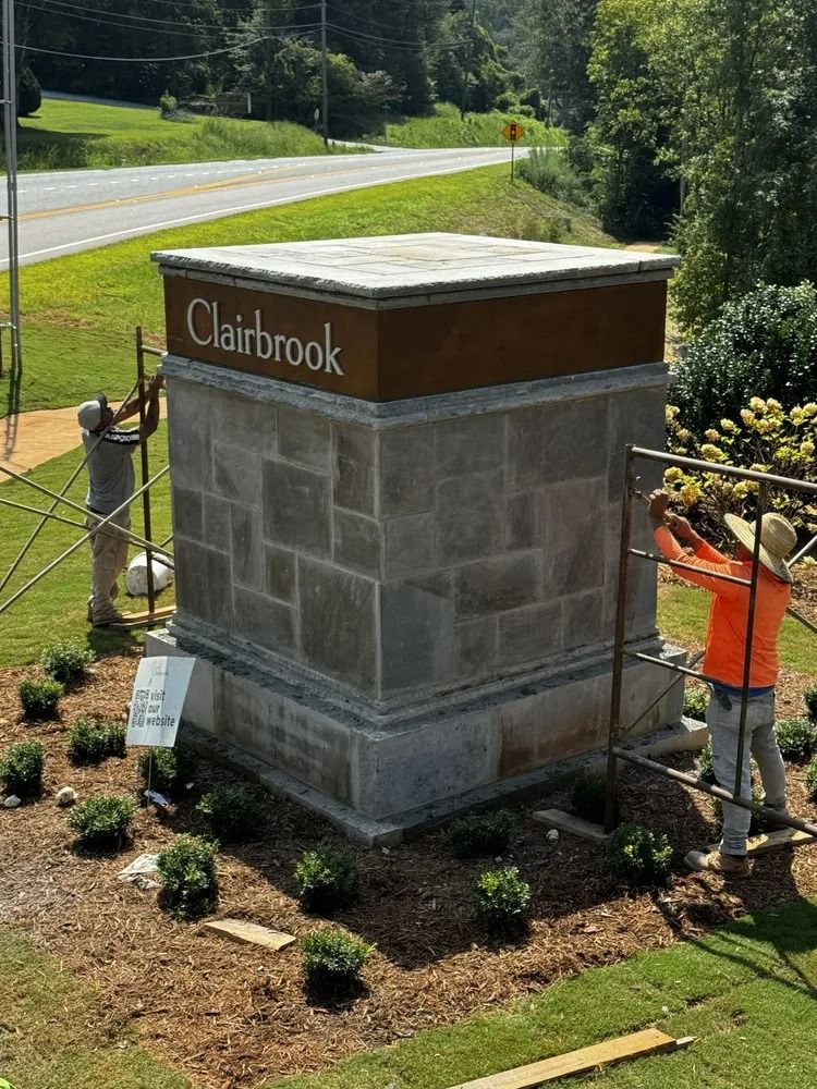 Stone monument sign reading “Clairbrook” beside a roadside, with a worker planting flowers nearby.