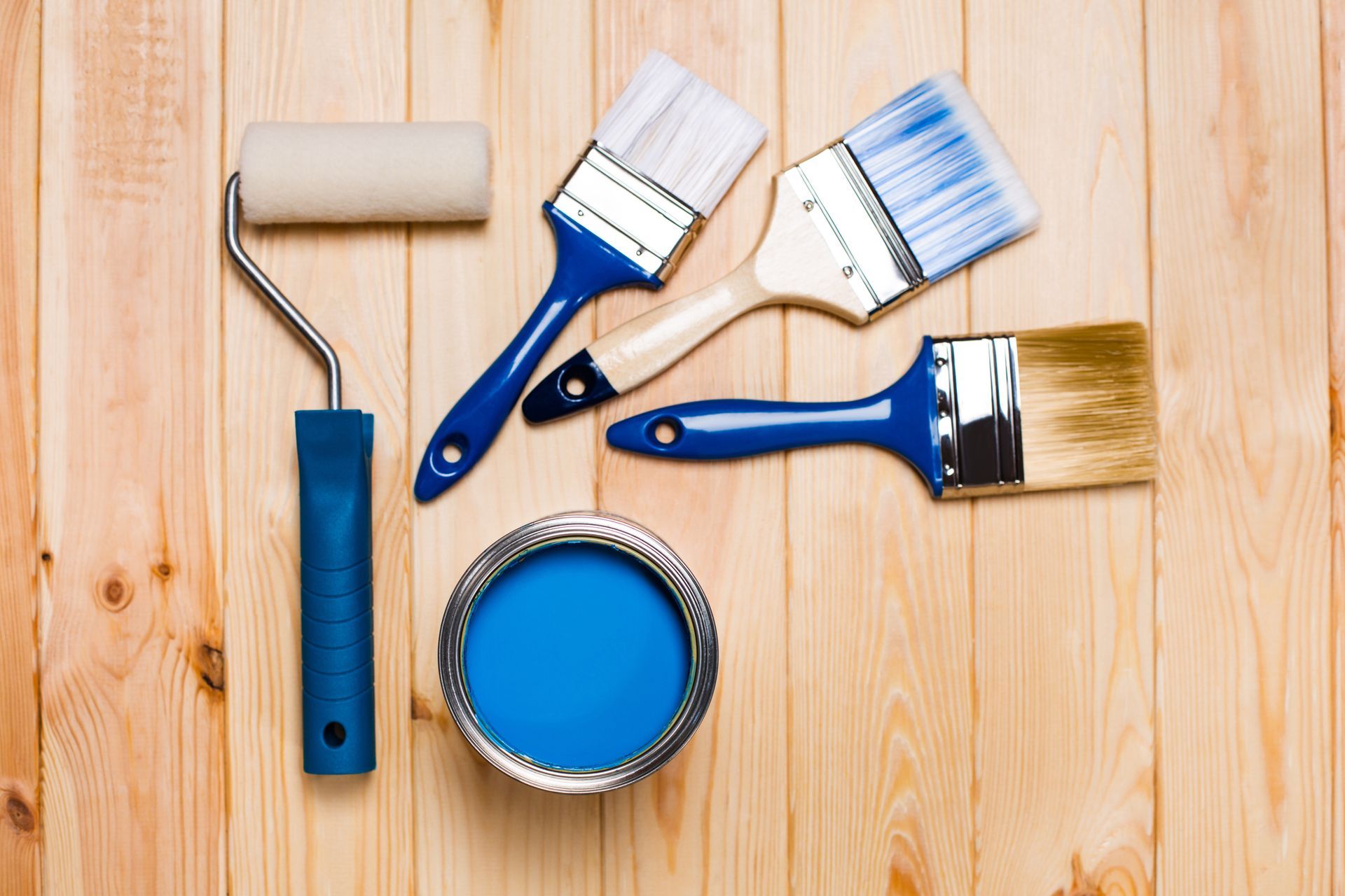 Paint brushes, roller, and blue paint can on wooden table, showcasing commercial paint supplies.
