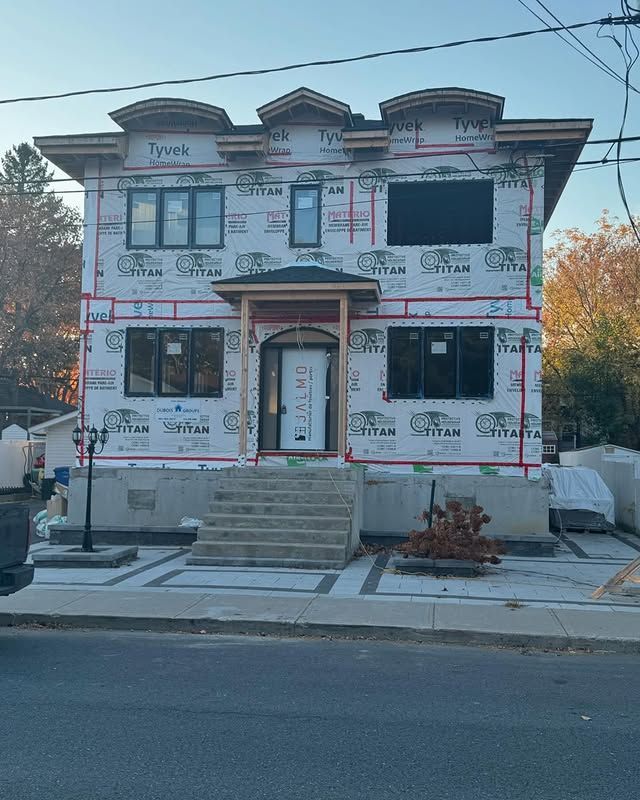 Maison à deux étages en construction, recouverte de Tyvek. L'entrée principale comporte un escalier, une porte et des fenêtres.