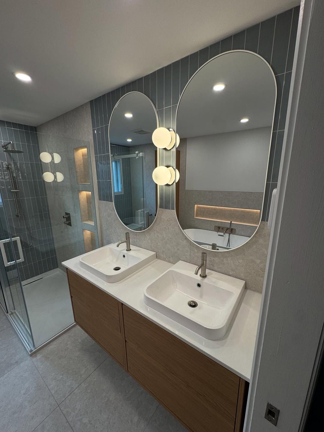 Salle de bains moderne avec double vasque, miroirs ovales et meubles en bois clair. Accents de carrelage gris et bleu.