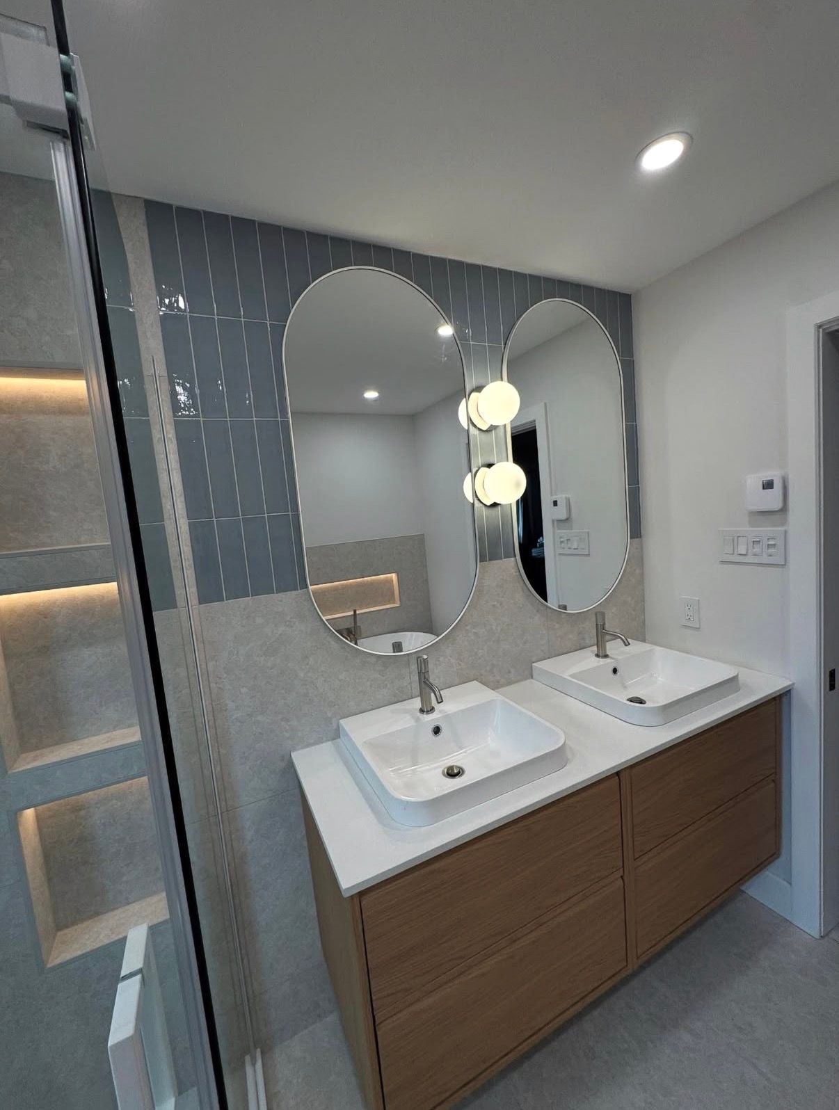 Salle de bains moderne avec miroirs ovales, lavabos blancs et meuble-lavabo en bois. Mur d'accent en carrelage bleu.