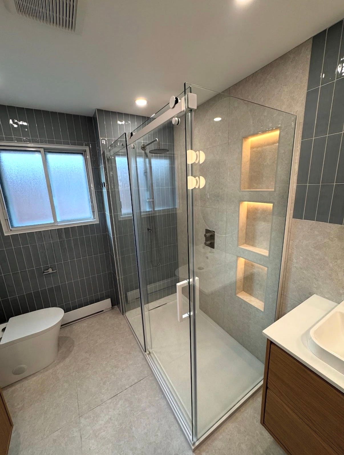 Salle de bains moderne avec douche en verre, carrelage gris et fenêtre.