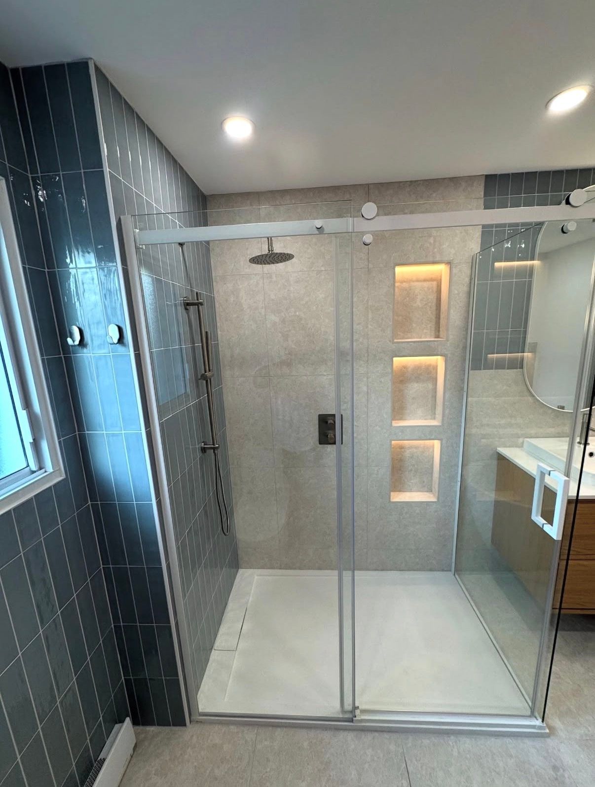 Salle de bains moderne avec paroi de douche en verre, carrelage gris et étagères intégrées.