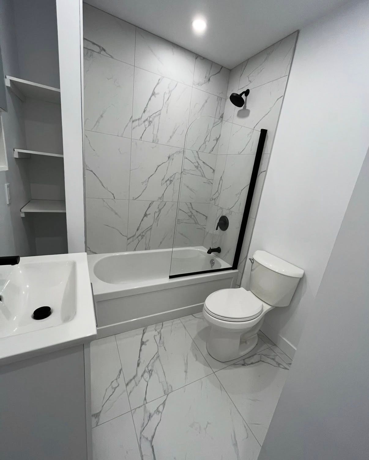 Salle de bains avec carrelage blanc imitation marbre, baignoire, toilettes et lavabo. Pommeau et cadre de douche noirs.