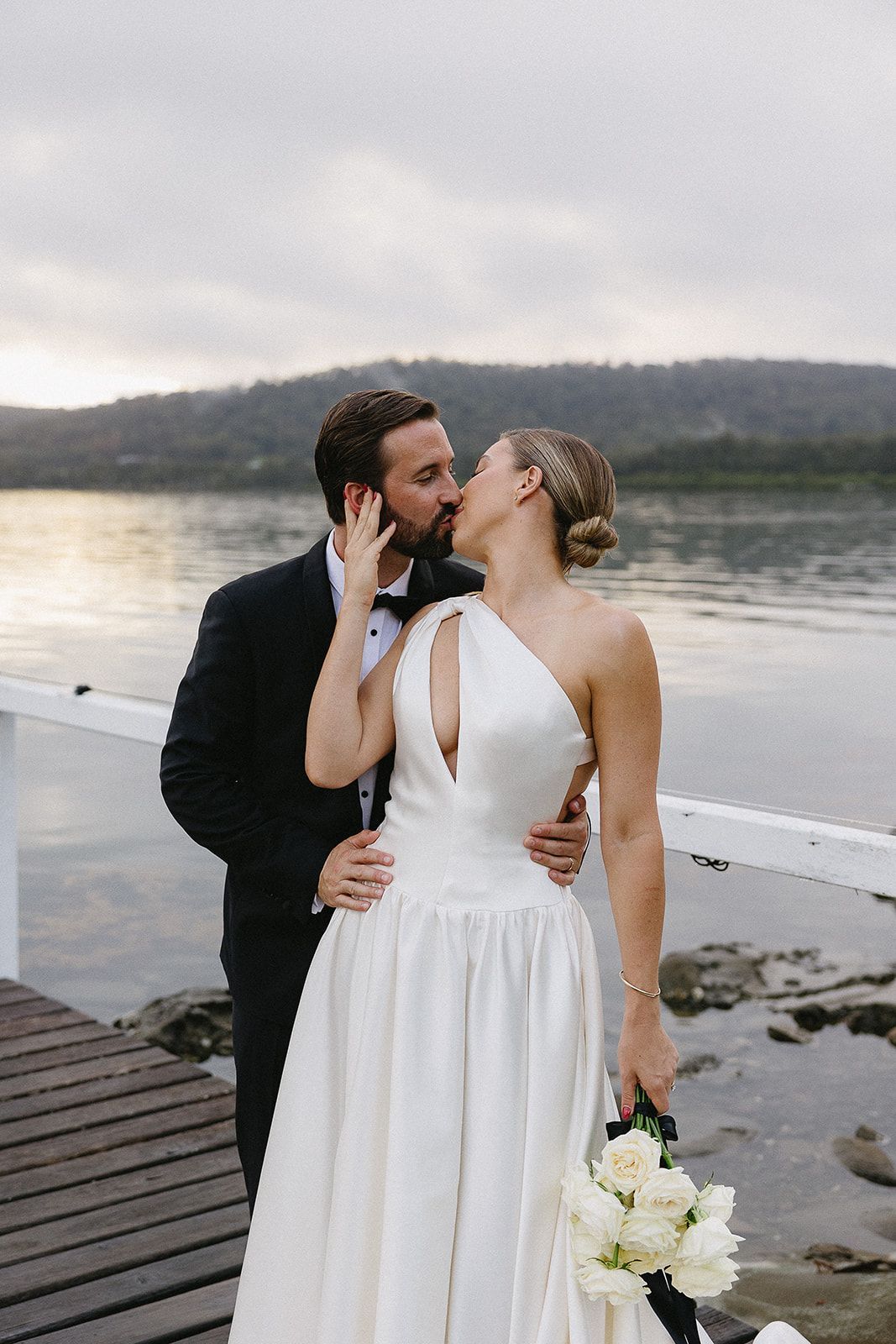 A bride and groom are kissing on a dock by the water.