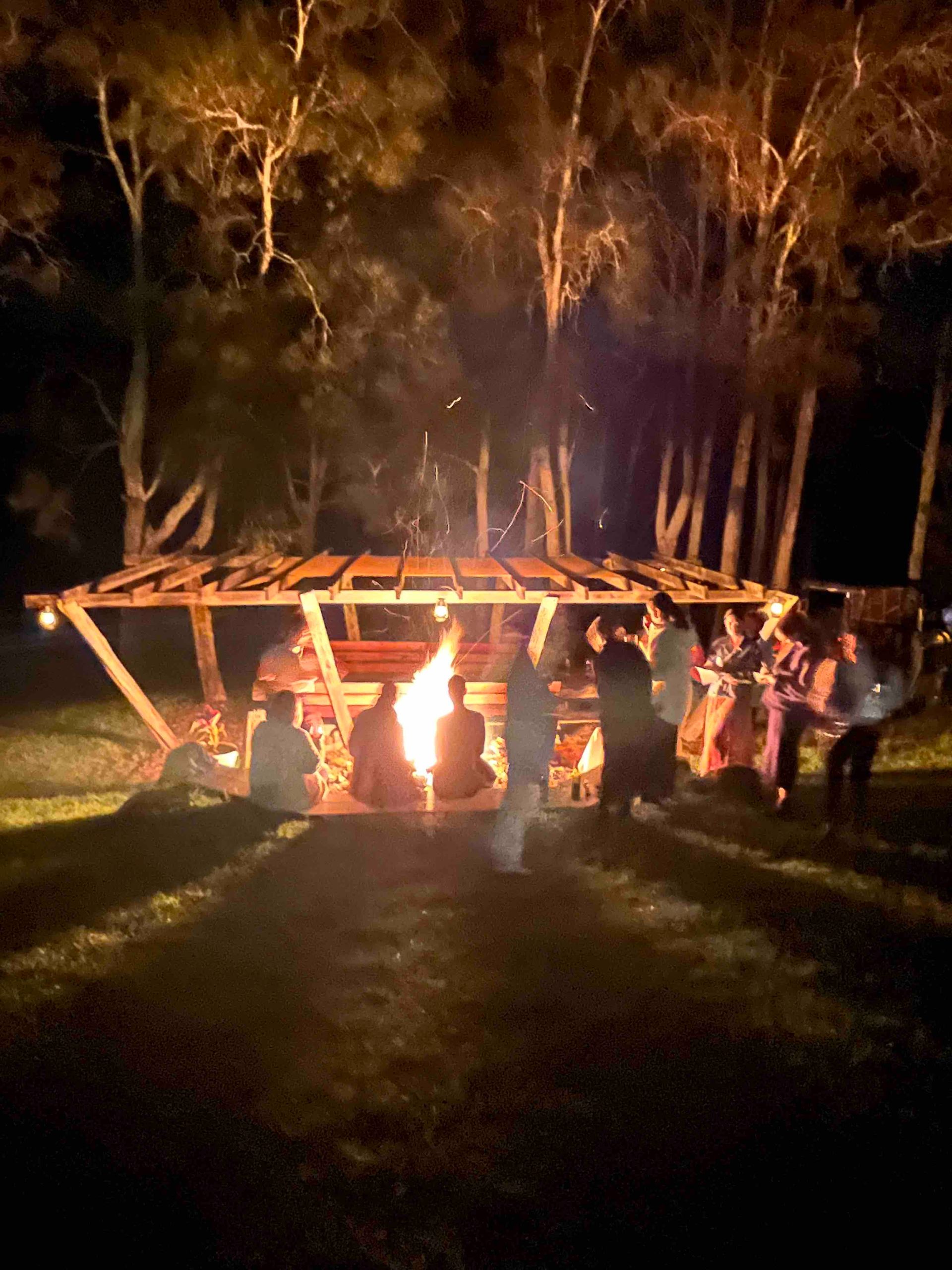 A group of people are standing around a fire pit at night.