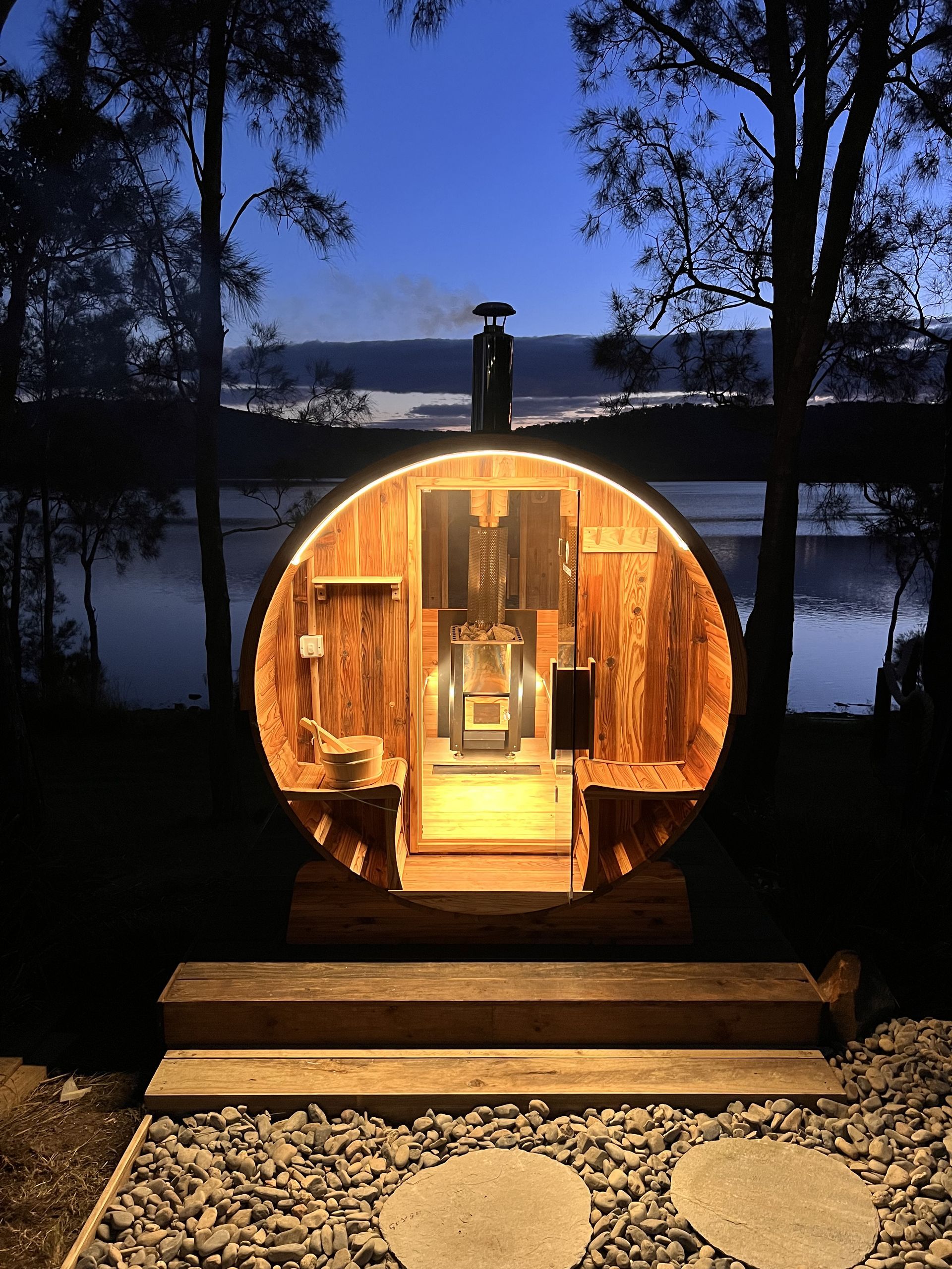 A wooden barrel sauna is lit up at night with a view of a lake.