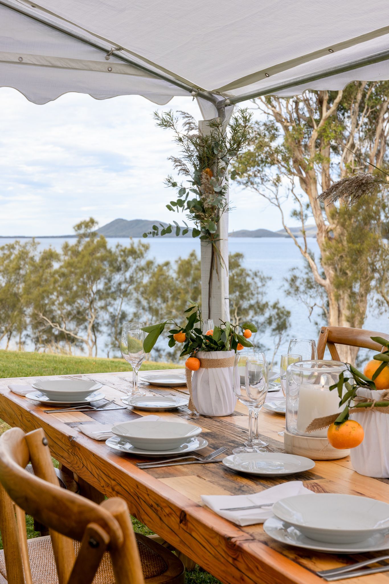 A long wooden table with plates , bowls and glasses under a tent overlooking the ocean.