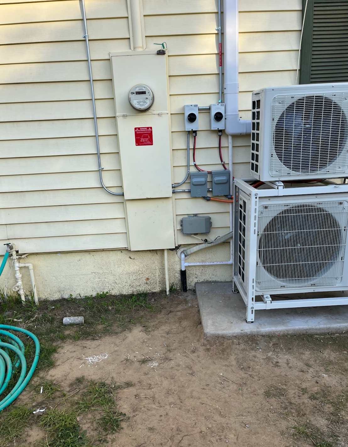 A gray residential air conditioning unit sits on a concrete pad against a red brick house near garden bushes.