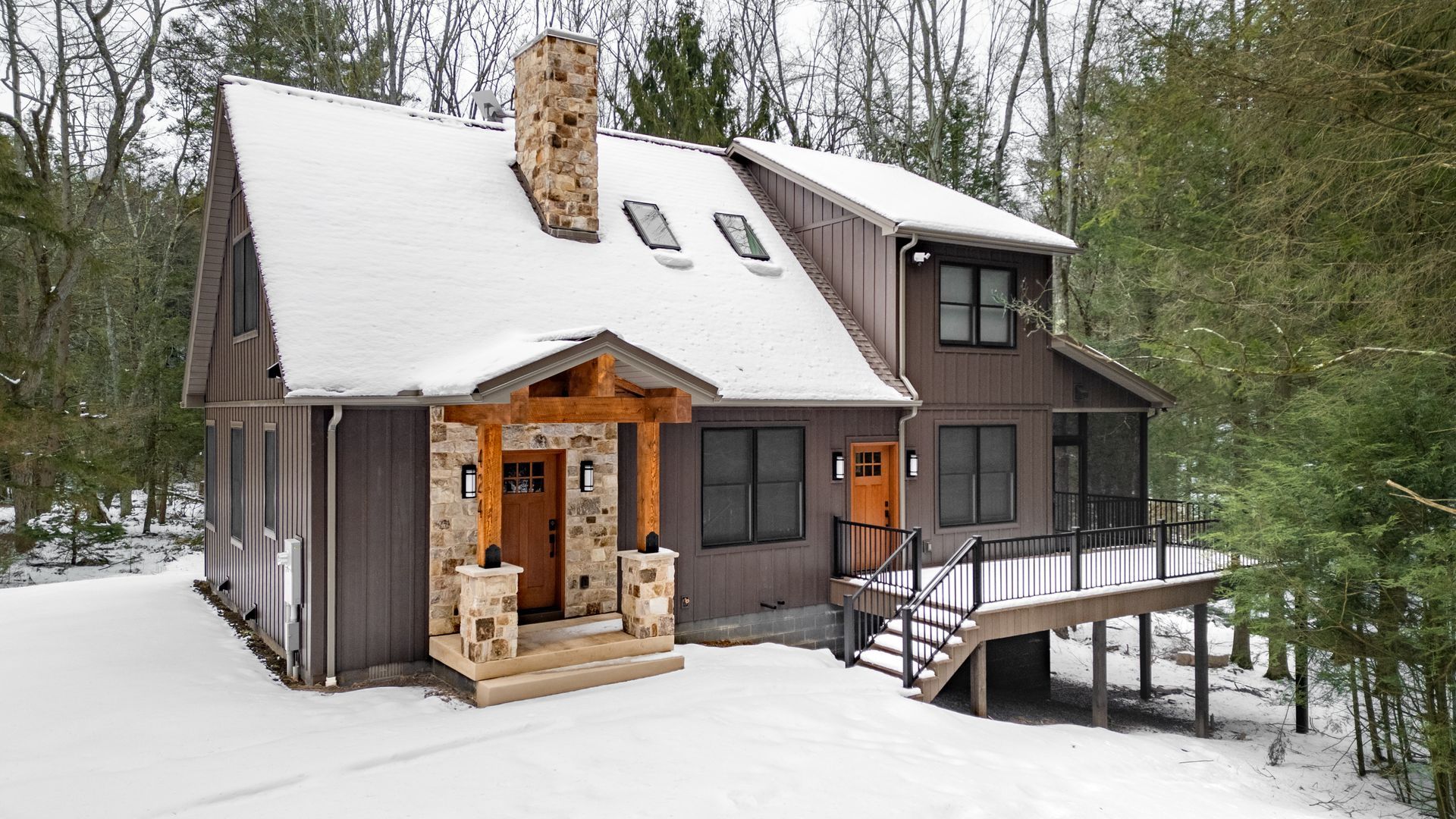 Snow-covered rustic cabin with wood siding, stone chimney, and a small deck in a wooded winter setting