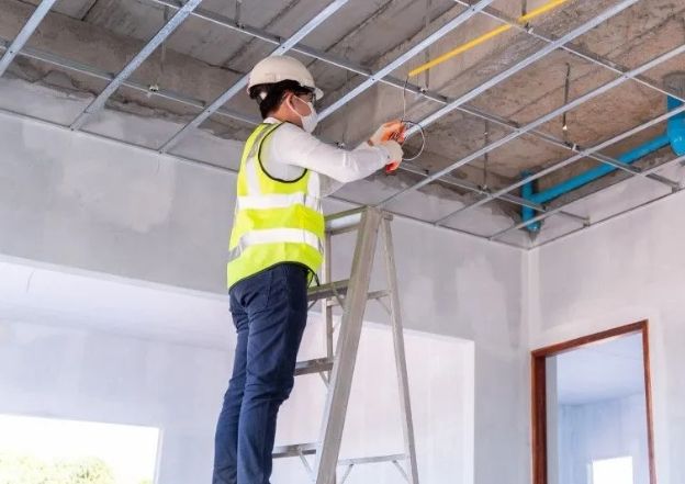 Technician on a ladder performing electrical maintenance on a ceiling grid.