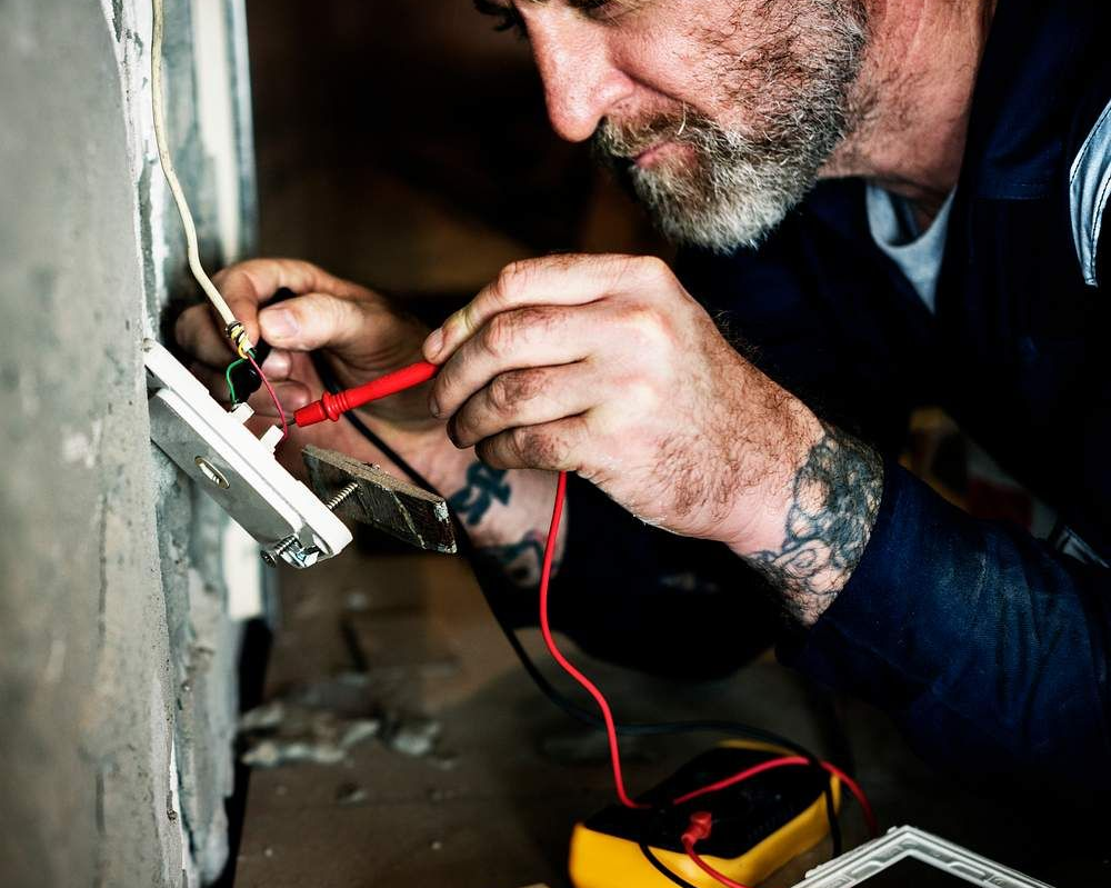 Electrician using a digital multimeter to troubleshoot a wall light switch.