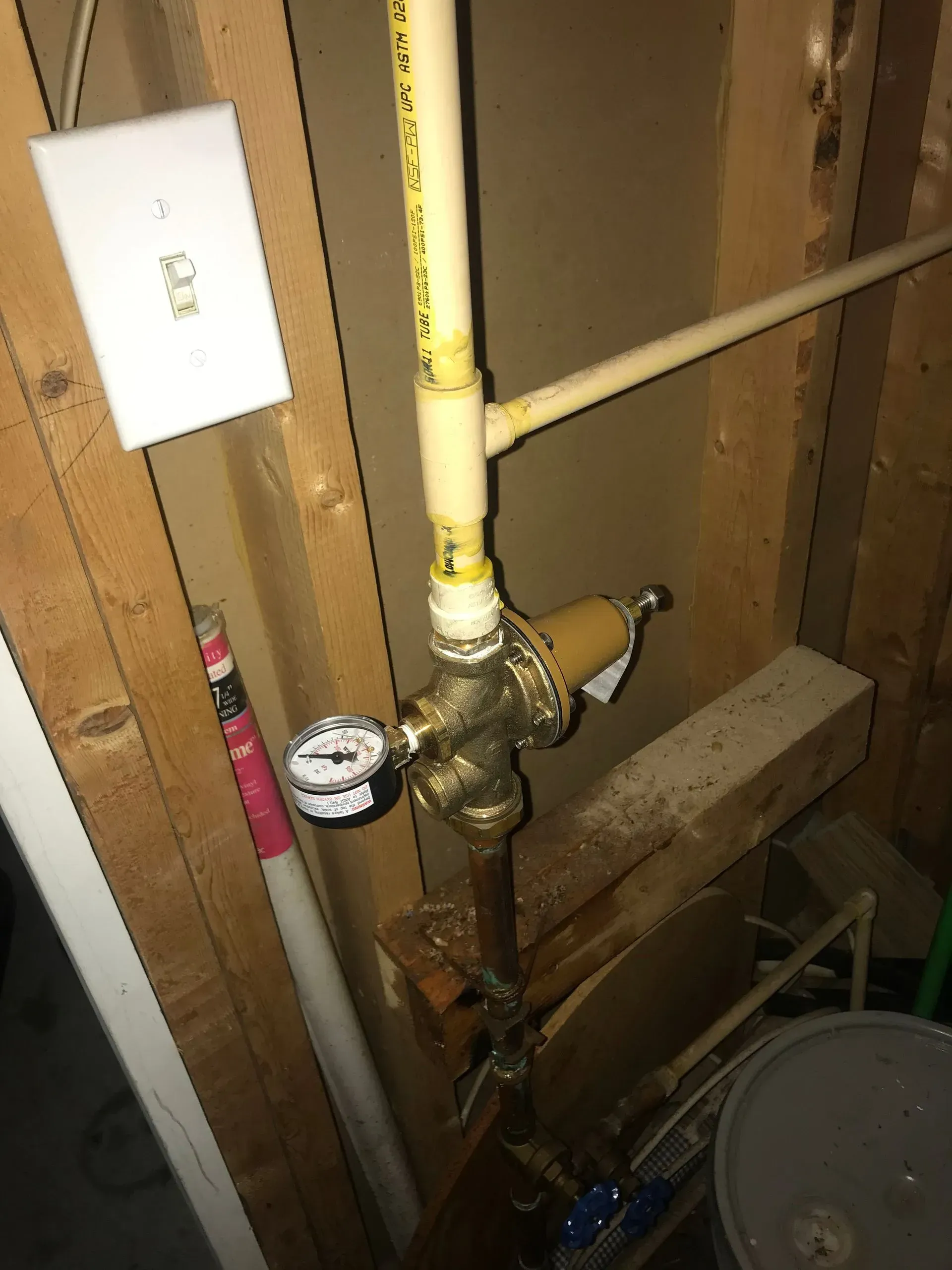 Water pressure regulator with gauge, connected to yellow PVC pipes in a basement setting.