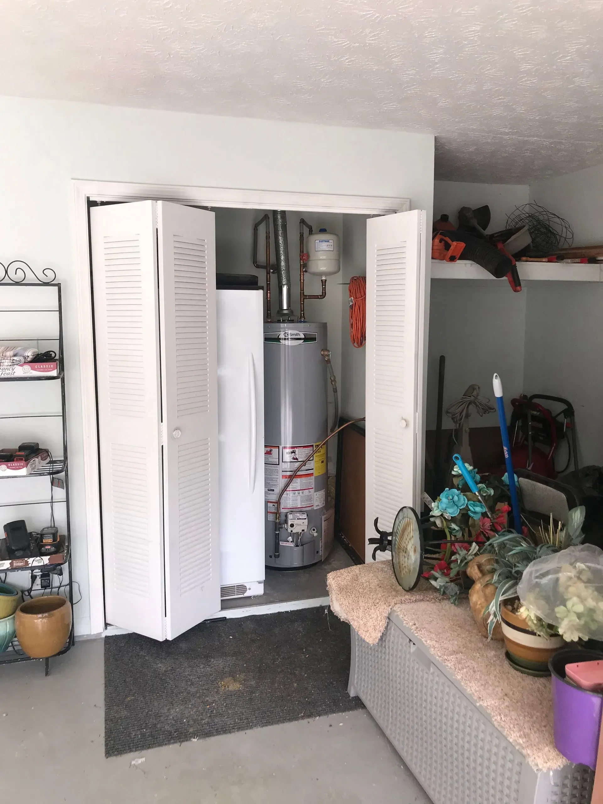 Water heater and refrigerator inside a closet with folding doors in a room. Various items stored nearby.