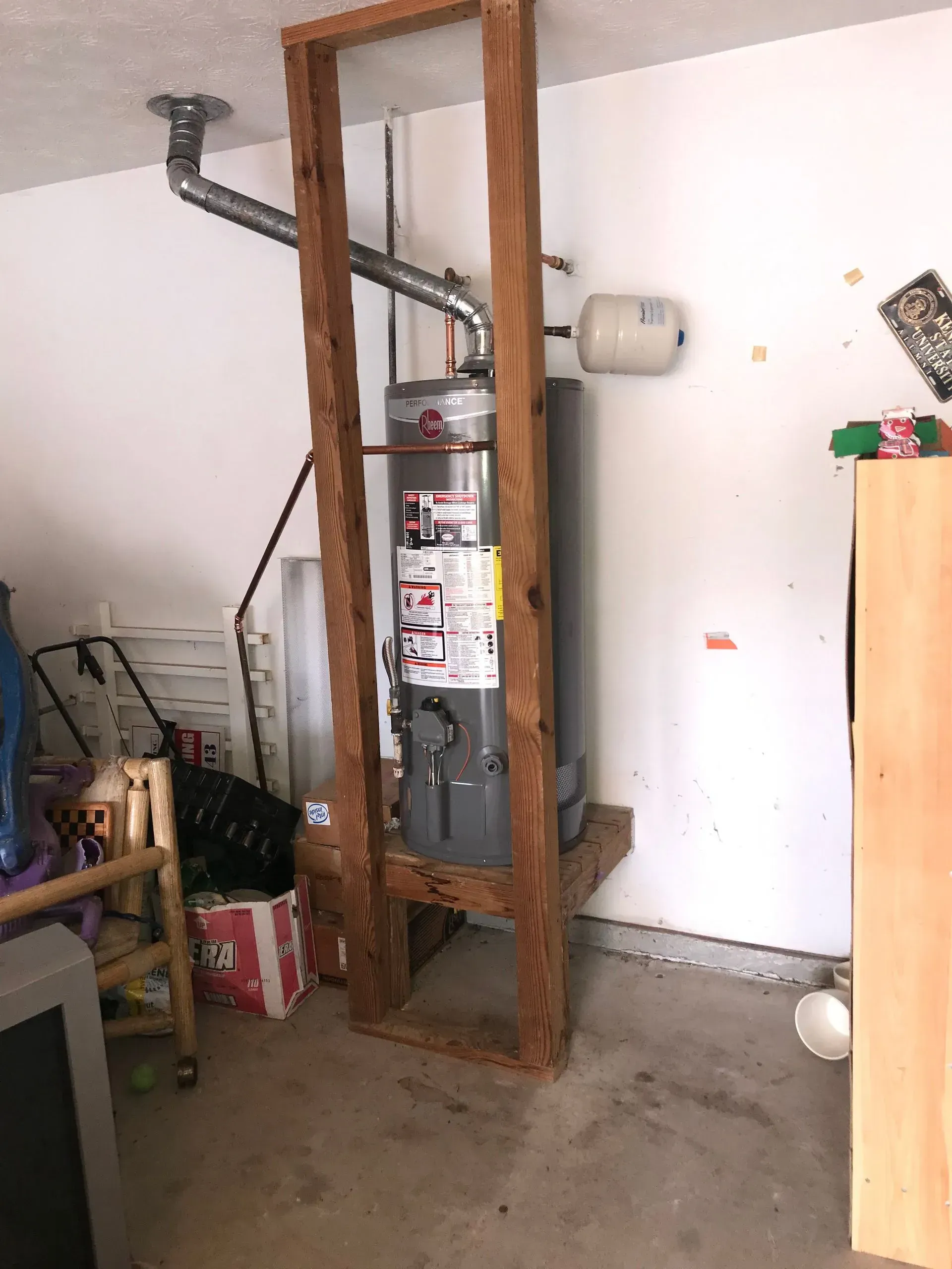 Water heater framed in a garage. The wooden frame appears DIY. Includes a tank and vent pipe.