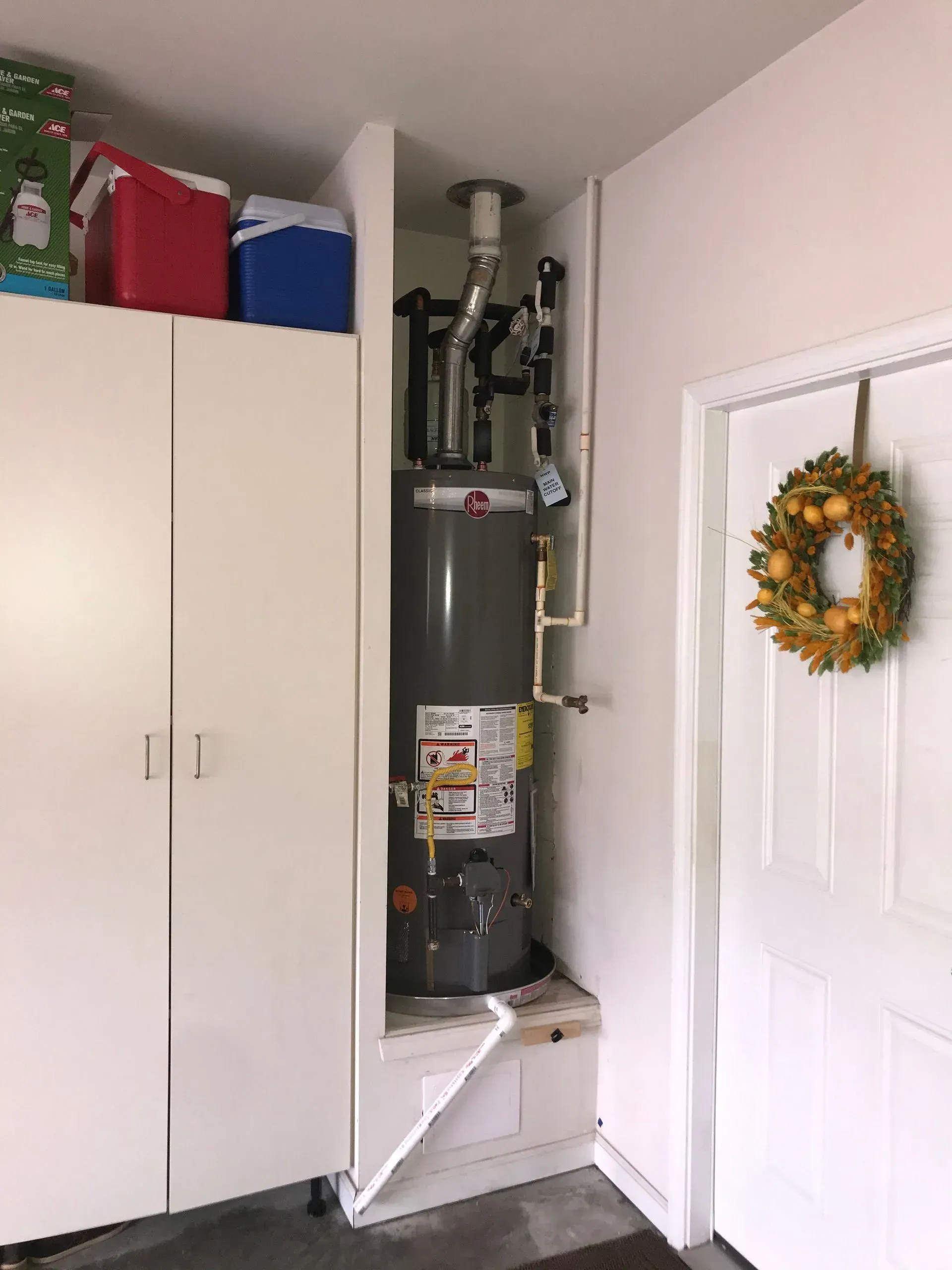 Water heater in a garage alcove next to a cabinet and door. A wreath hangs on the door.
