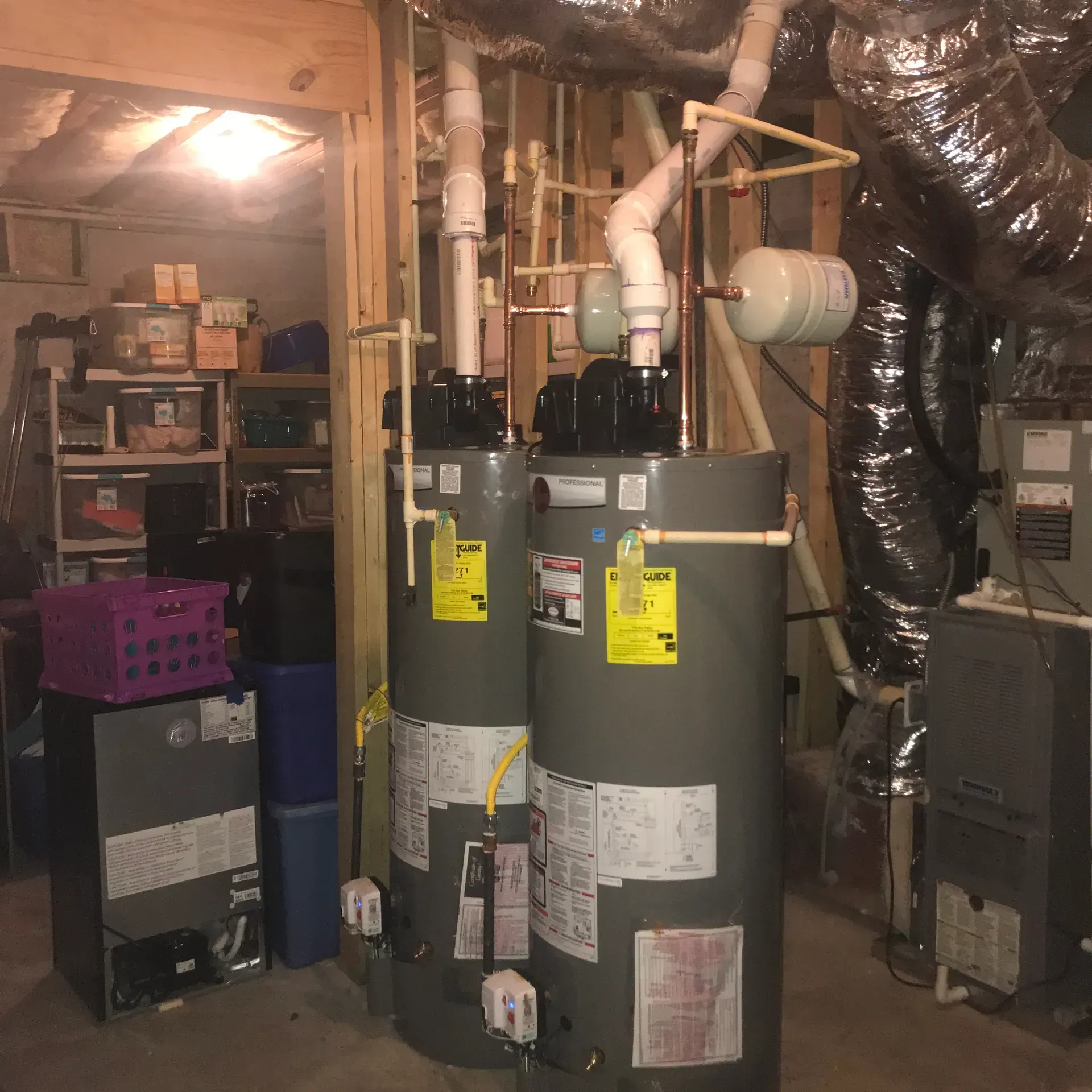 Two water heaters, with copper pipes, in a basement next to a furnace and storage shelves.