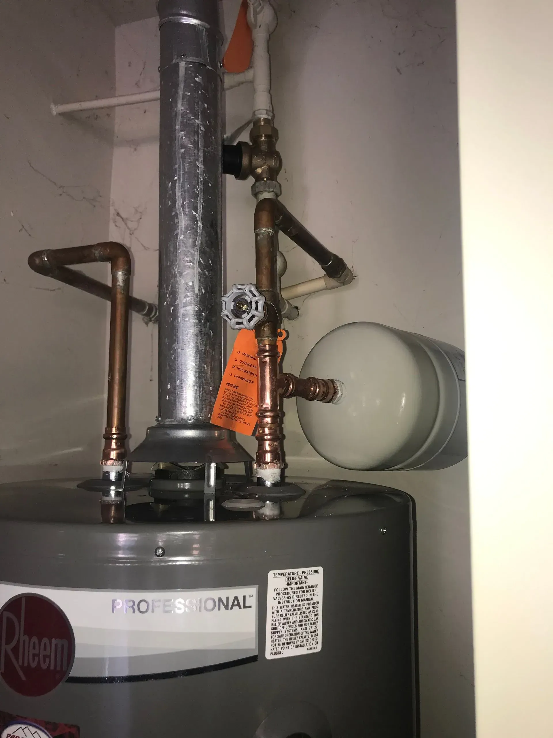 A Rheem water heater in a utility closet with copper pipes, a white expansion tank, and a silver exhaust pipe.