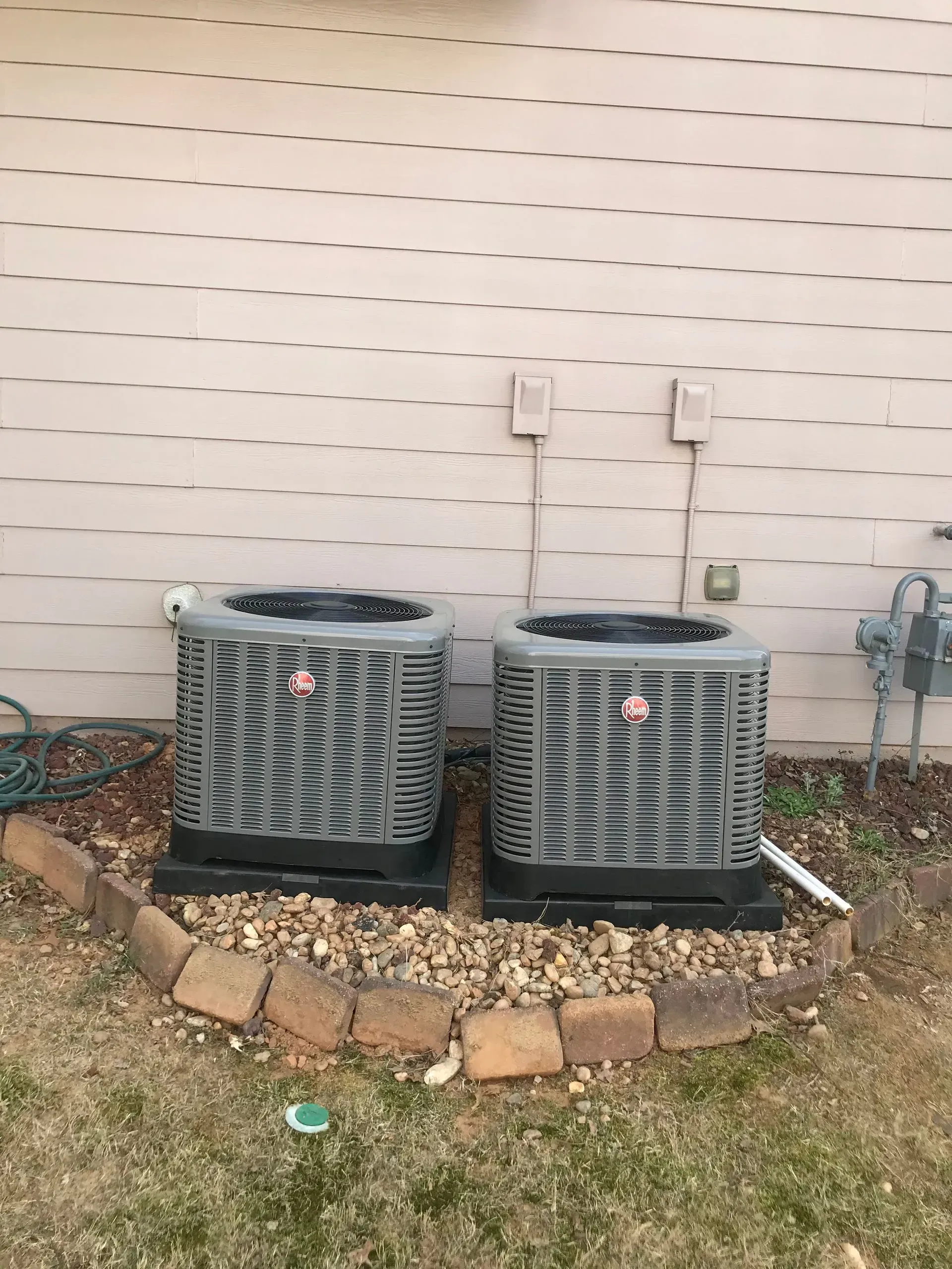 Two grey air conditioning units side-by-side outside a house with beige siding, surrounded by landscaping stones.