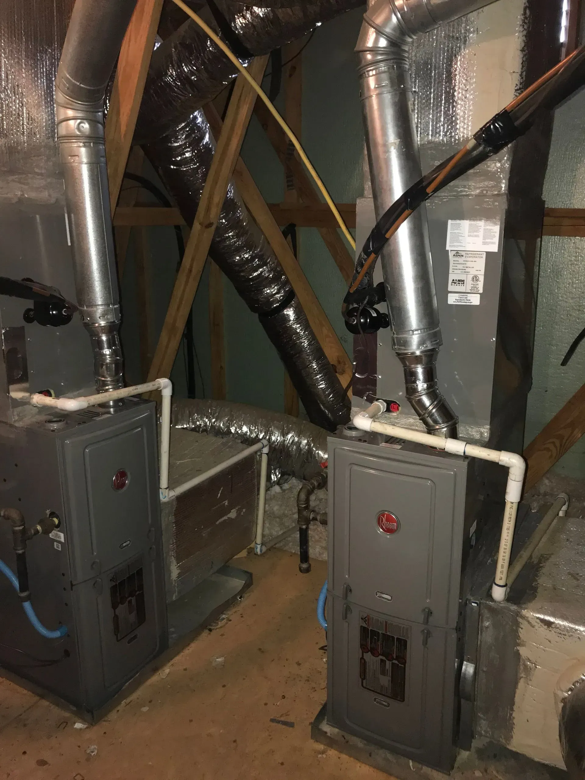 Two gray Rheem HVAC units in an attic, with ductwork and wooden supports.