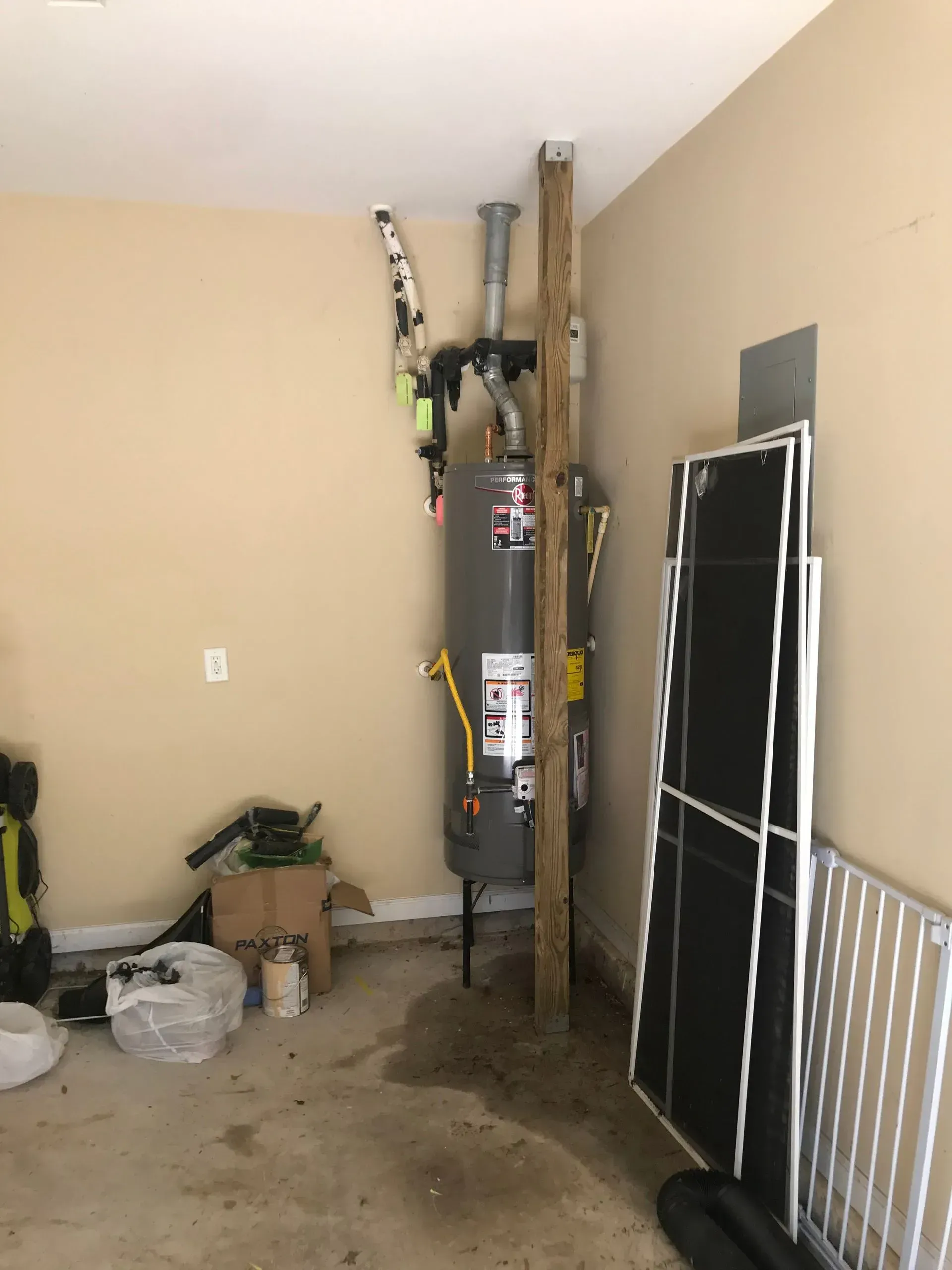 Water heater in a garage, with wood support beams, concrete floor, and various items stored nearby.